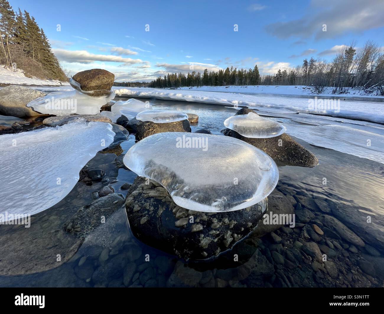 Flussfelsen mit durchscheinenden Eiskappen entlang eines halbgefrorenen Flusses mit blauem Himmel und Bäumen entlang der Küste - Smartphone-aufgenommenes Stockfoto