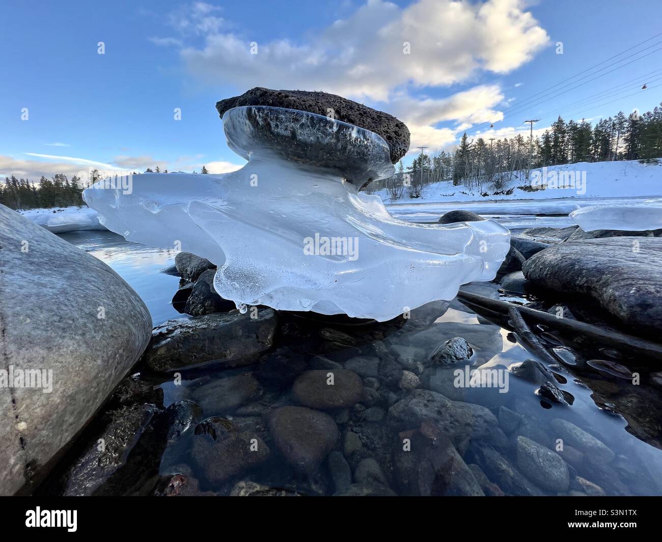 Flussfelsen auf einer durchscheinenden Eisskulptur entlang eines halbgefrorenen Flusses mit blauem Himmel und Bäumen am Ufer - Smartphone-aufgenommenes Stockfoto