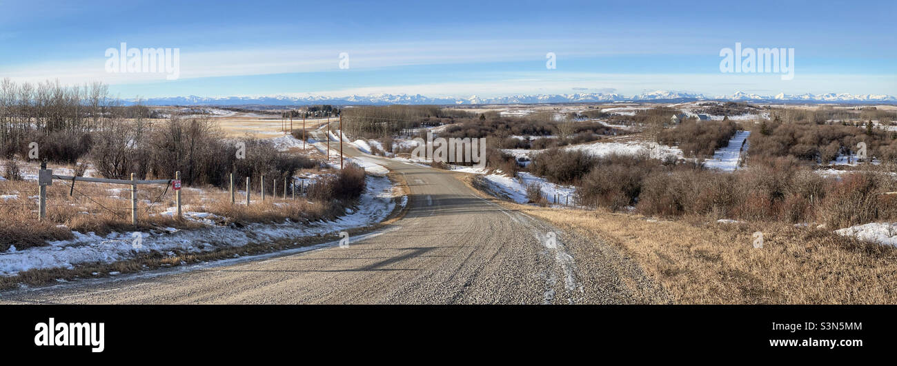 Panorama einer Landschaft am Fuße der Berge, in der Nähe von Calgary, Alberta, Kanada. Eine kurvenreiche Straße führt den Hang hinunter, mit Bäumen auf beiden Seiten, in Richtung der entfernten kanadischen Rocky Mountains. Stockfoto