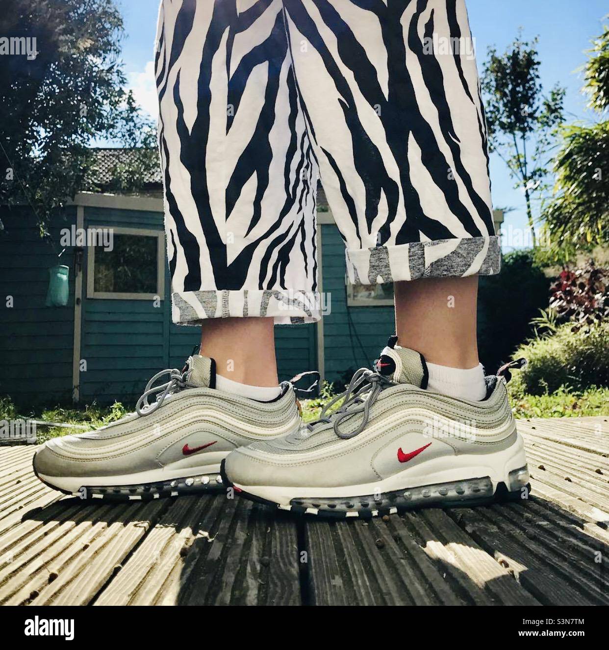 Beine mit Zebramuster – eine Frau im Zebra bedruckte dreiviertel lange Hose und Nike Sportschuhe, die auf der Terrasse im Garten stehen Stockfoto