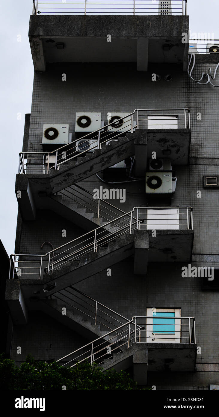 Das mehrstöckige Gebäude verfügt über eine Feuertreppe mit eisernen Geländern, blauen Türen und Klimaanlage. Stockfoto