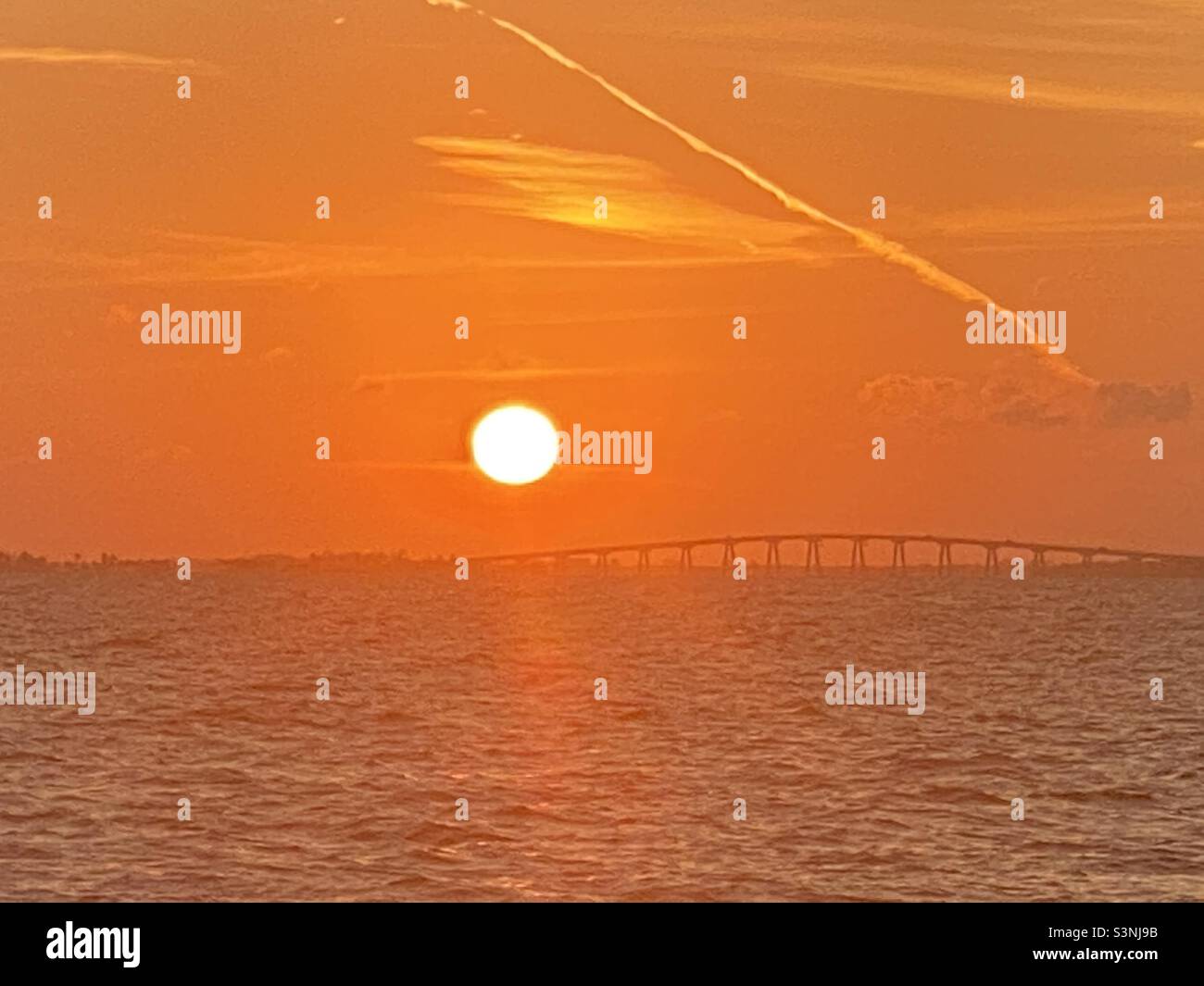 Strahlender orangefarbener Himmel mit der riesigen Sonne, die über eine Brücke und Küste in FT Myers Beach, Florida, untergeht, Blick von einem Boot auf dem Wasser, Silhouette Sonnenuntergang Stockfoto