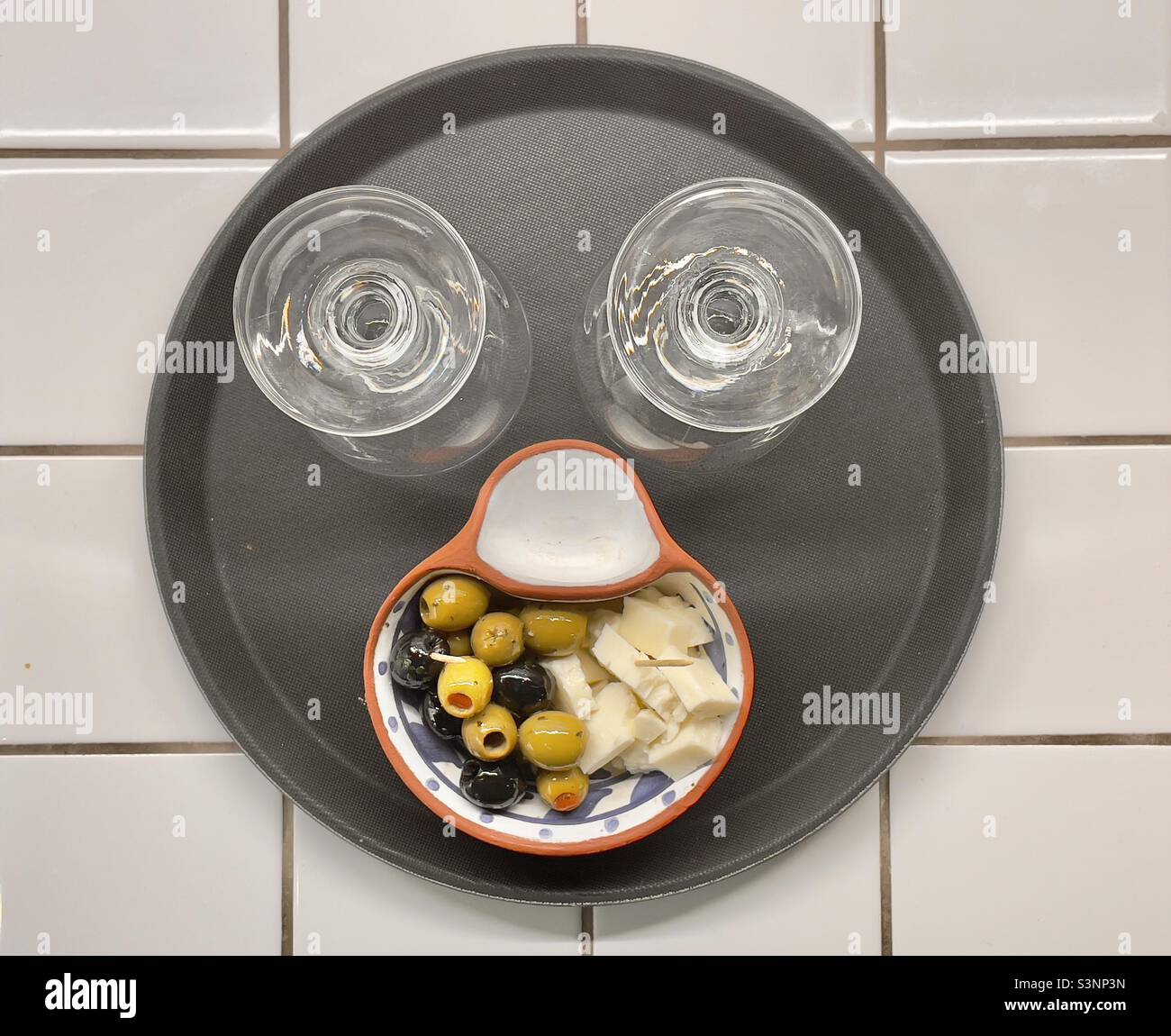 Ein schwarzes Tablett mit zwei Weingläsern und einem Gericht aus Oliven, das zu einem humorvollen Gesicht arrangiert wurde Stockfoto