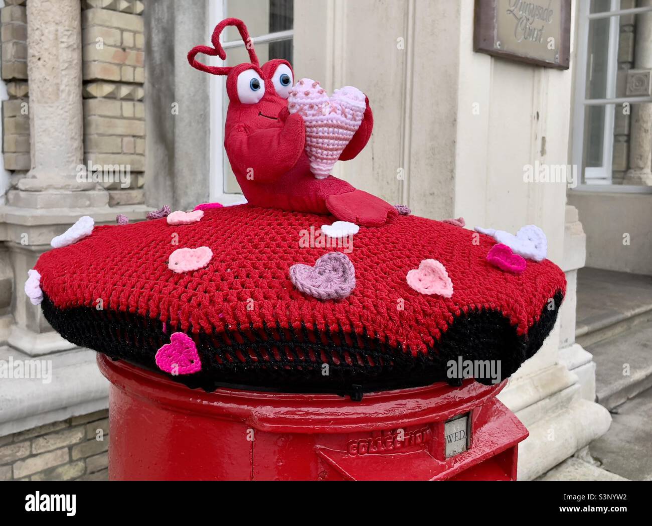 Ein britischer Briefkasten mit Gestricktem Wollhut mit Motiven zum Valentinstag, eine charmante, skurrpische Szene im Hafen von Harwich - Smartphone-aufgenommenes Stockfoto