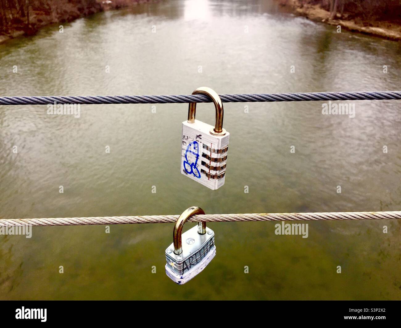 Der Fluss der Liebe. Liebesschloss mit Fischmotiv. Zwei Schlösser an Stahlseilen mit Blick auf das fließende Wasser. Frisch. Noch kein Rost. Klassische Symbole. - Smartphone-aufgenommenes Stockfoto