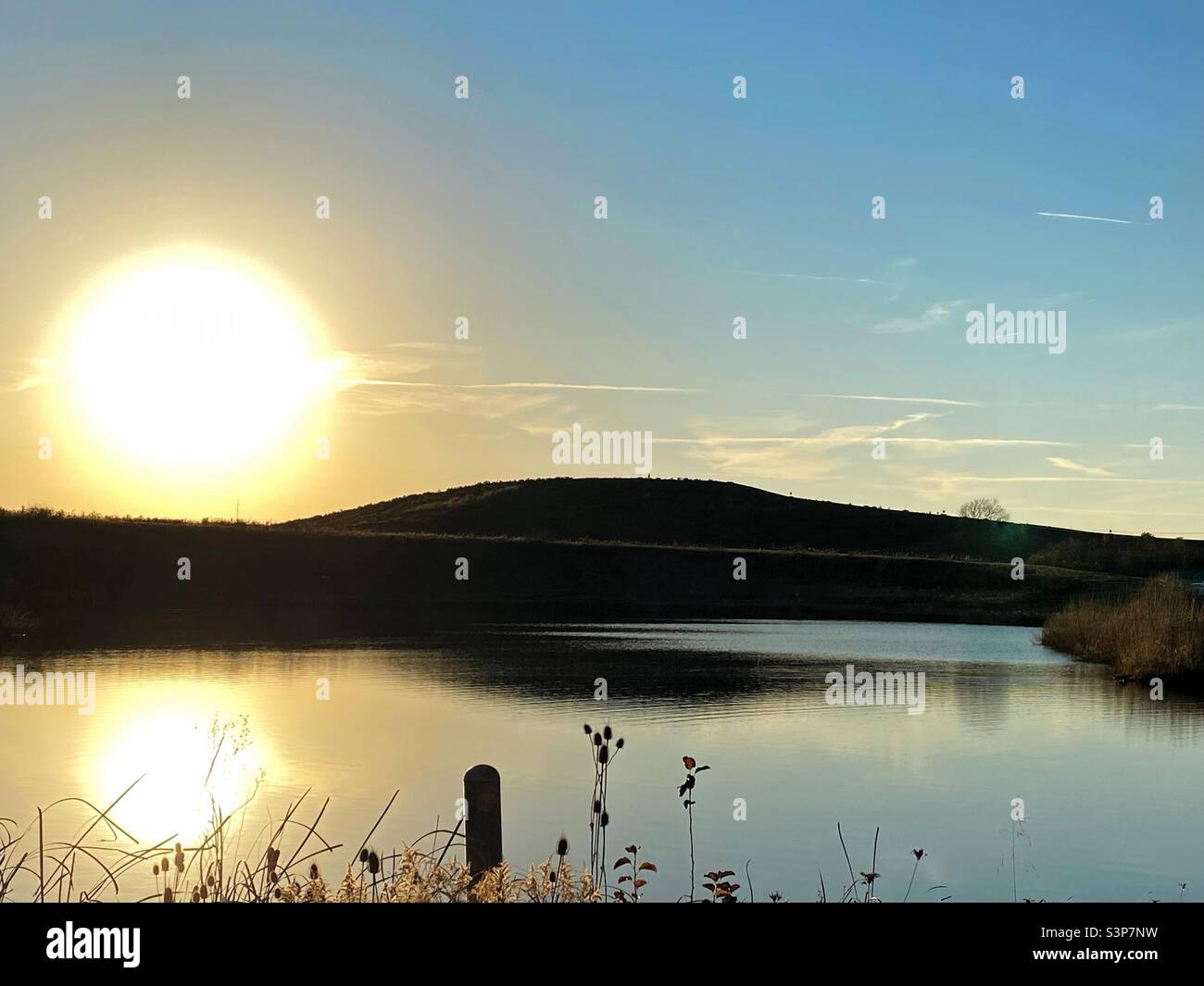 Die Sonne beginnt hinter einem Hügel unterzugehen. Die Spiegelung der Sonne kann im See im Vordergrund gesehen werden. Stockfoto