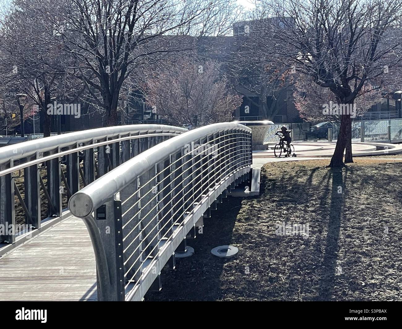 Eine leere Grünfläche in Salt Lake City, Utah, USA, die über eine Brücke in Richtung eines kleinen, familienorientierten Skulpturenparks blickt. In der Ferne kann man eine Einzelstatue eines kleinen Jungen sehen, der mit dem Fahrrad unterwegs ist. - Smartphone-aufgenommenes Stockfoto