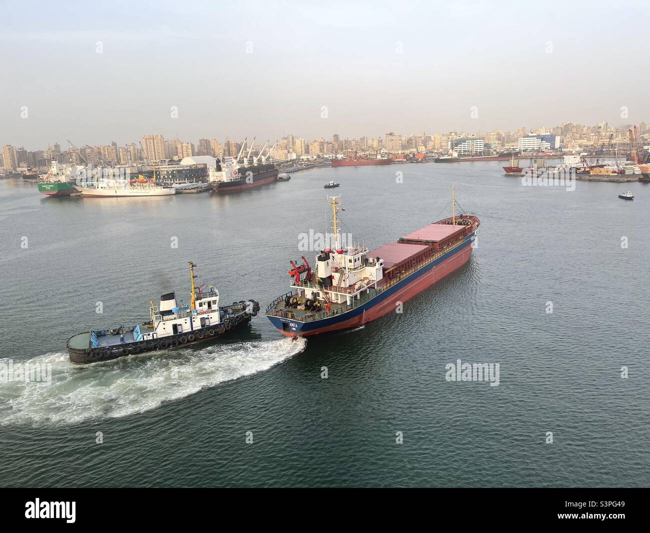 Kleines Bulk-Carrier-Schiff durch Schlepper während der Annäherung Liegeplatz im Hafen von Alexandria, Ägypten unterstützt. Im Hintergrund sind der Bau der Stadt und andere festfahrende Schiffe während des Frachtbetriebs. Stockfoto