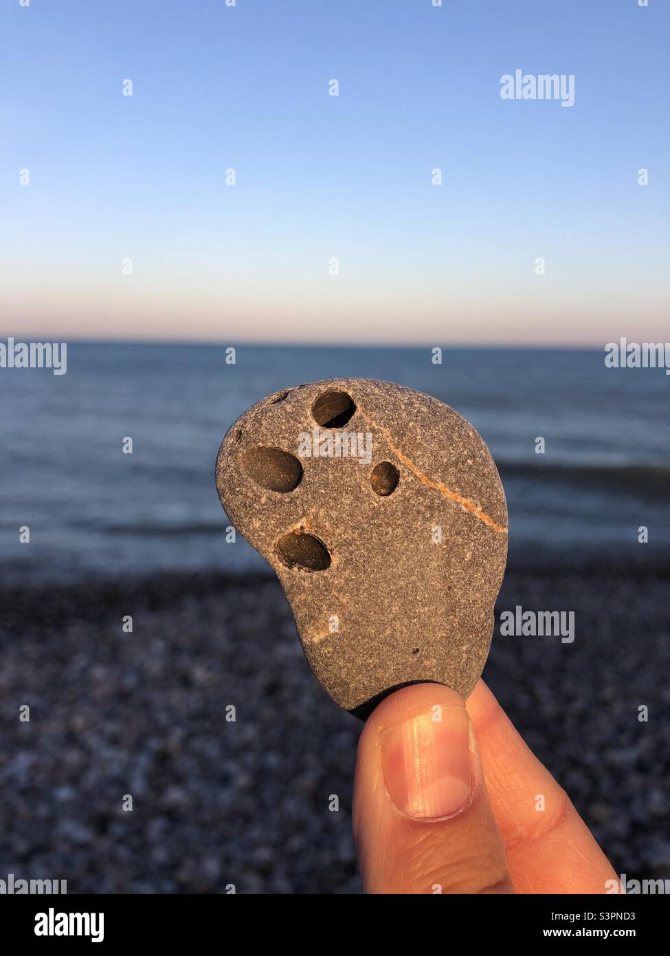 Stein mit vier Löchern, der im Abendlicht am Meer in der Hand gehalten wird - Smartphone-aufgenommenes Stockfoto