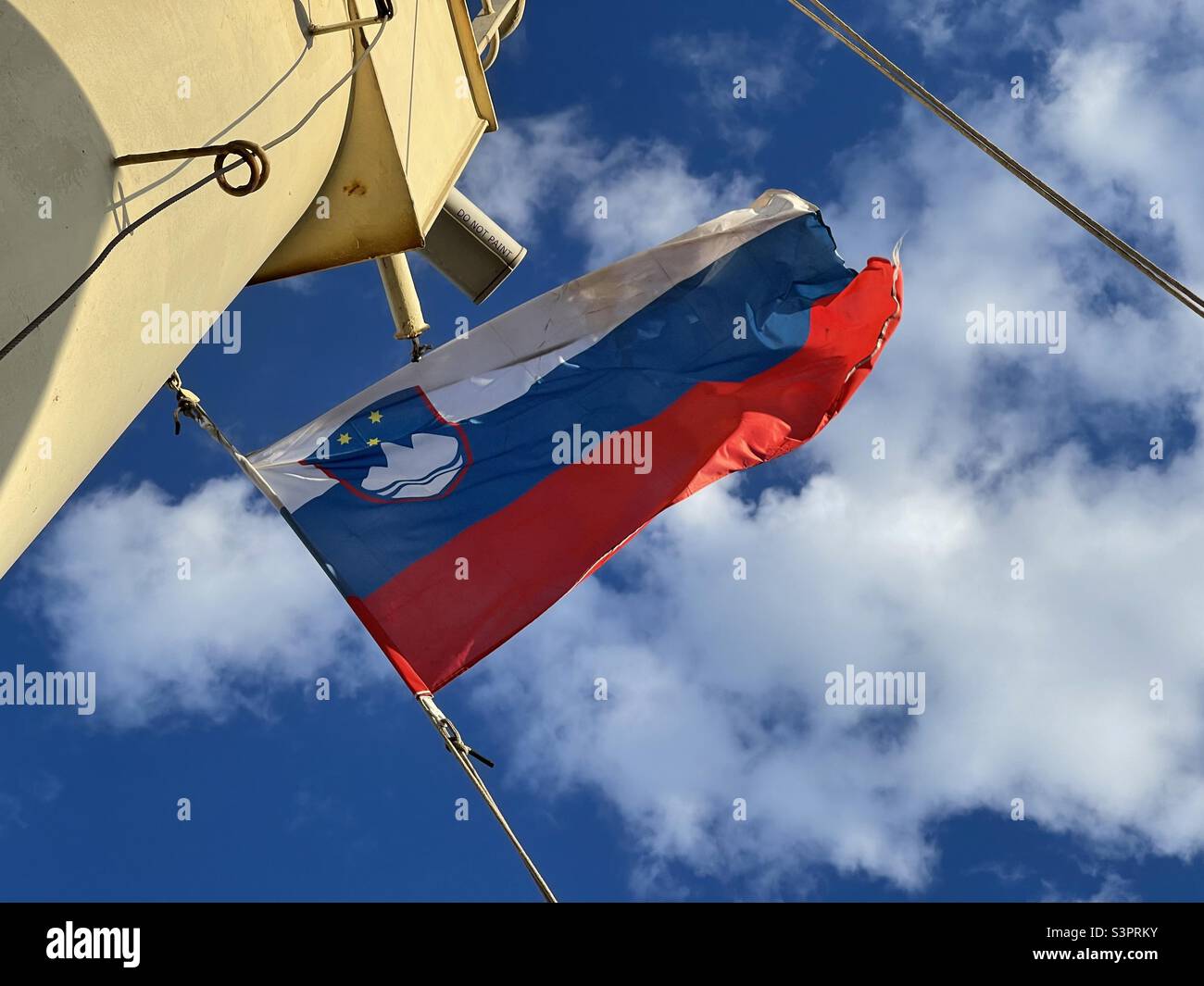 Slowenische Flagge auf dem Navigationsmast des Frachtschiffes, das im slowenischen Hafen bei windigem, sonnigem Frühlingswetter festgemacht wurde. Auf dem Hintergrund blauer Himmel und weiße Wolken. Stockfoto