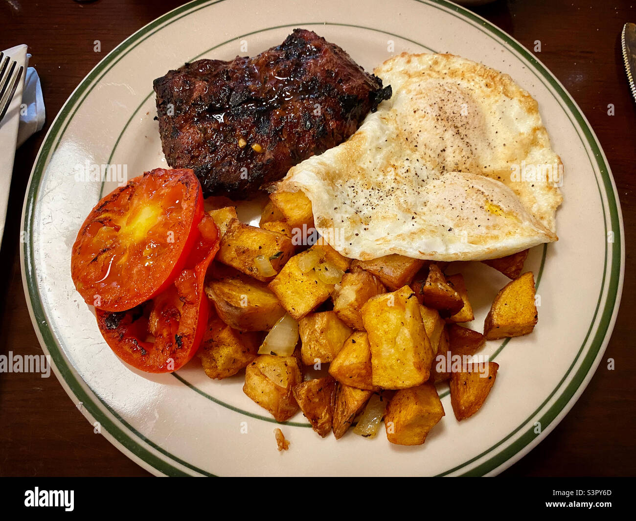 Frühstück mit Steak, Spiegeleiern, gebratenen Tomaten und Bratkartoffeln auf einem weißen Teller - Smartphone-aufgenommenes Stockfoto