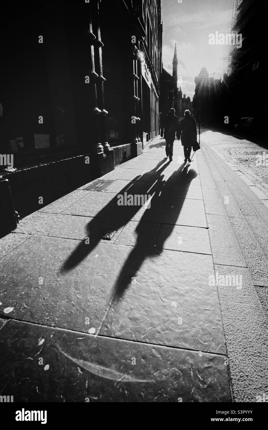 Ein Paar, das entlang der Royal Mile in Edinburgh geht, wird von der Nachmittagssonne beleuchtet und wirft lange Schatten Stockfoto