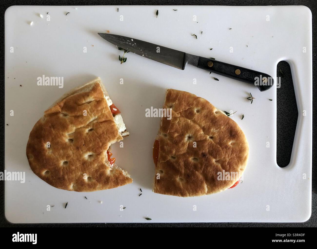Geröstete Focaccia auf einem Schneidebrett mit Kirschtomate, Feta-Käse, Thymian, Knoblauch und Messer - Smartphone-aufgenommenes Stockfoto