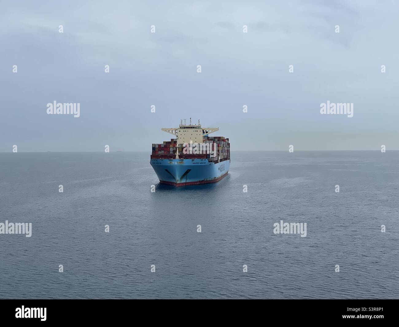 Voll beladenes riesiges Containerschiff mit blauem Rumpf von der Reederei Maersk, das bei regnerischem Wetter auf ruhigem Meer vor Anker liegt. Stockfoto