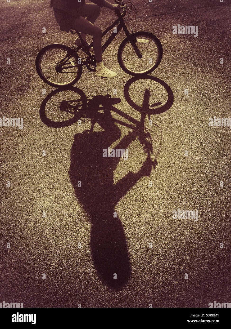 Die Sonne am späten Nachmittag wirft einen interessanten Schatten eines Kindes, das auf dem Fahrrad fährt. Schatten von Fahrrad und Fahrer, Foto ©️ COLIN HOSKINS. Stockfoto