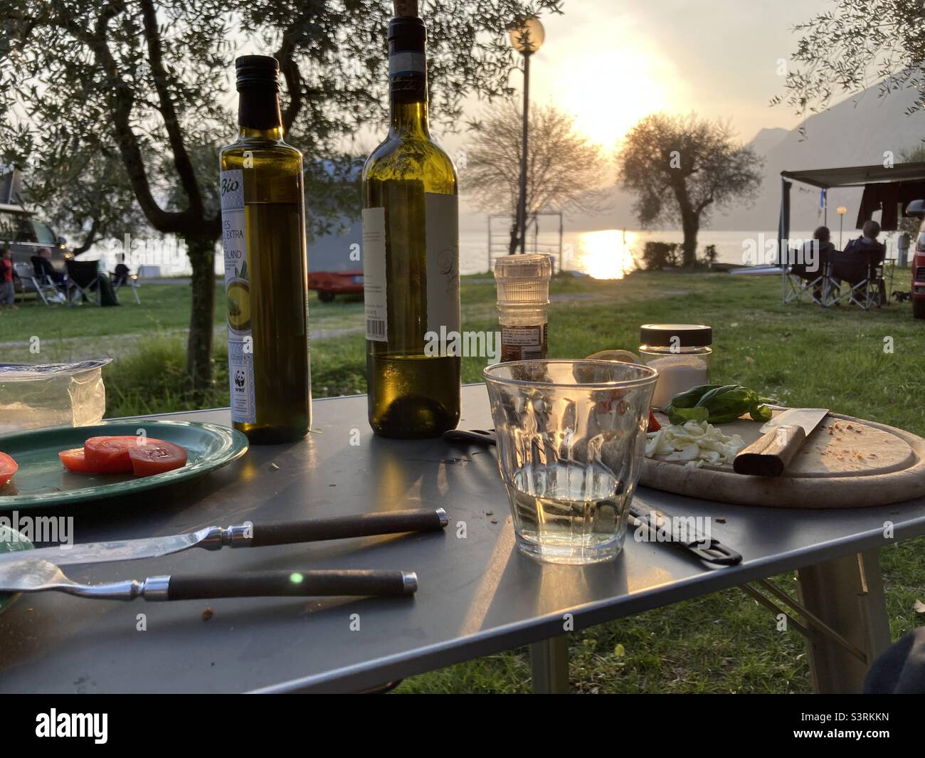 Ein Campingtisch mit Gläsern, einer Flasche Wein, Besteck, einem Schneidebrard, Olivenöl und Tellern mit Essensresten neben dem Lago d‘Iseo bei Sonnenuntergang, Iseo, Italien Stockfoto