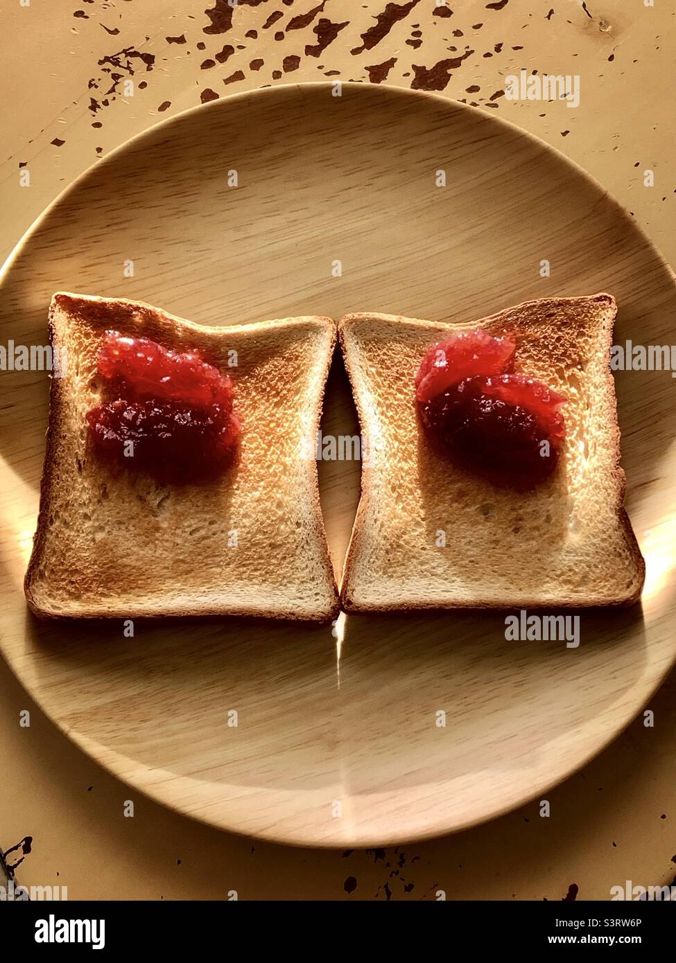 Zwei Toasts auf dem gelblichen Teller mit Erdbeerkonfitüren auf den Toasts wie Augen und sie befinden sich auf dem gelblichen Tisch und auch unter dem Morgenlicht vom Fenstervorhang. Stockfoto