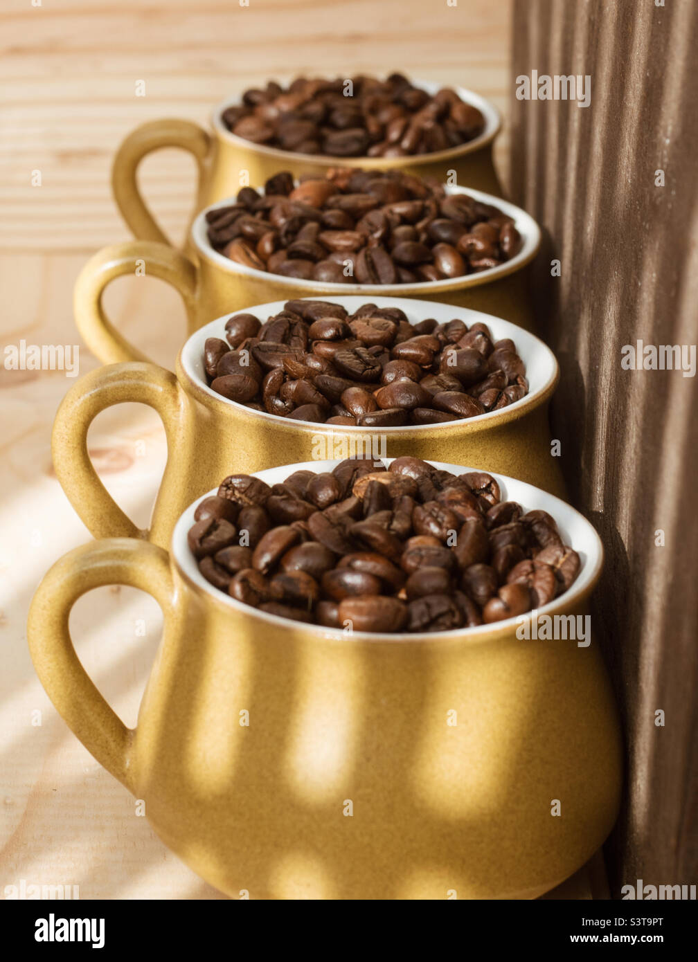 Eine Reihe von 4 Kaffeetassen, gefüllt mit Kaffeebohnen vor einem Holzhintergrund und gespaltenen Lichtschatten - Smartphone-aufgenommenes Stockfoto