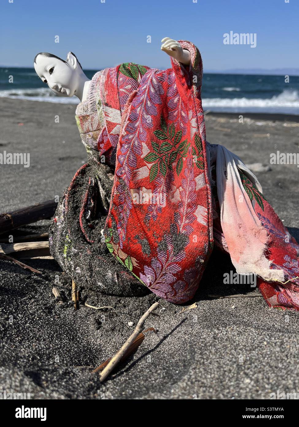 Eine japanische Puppe mit rotem Kimono, die in Strandsand vergraben ist - Smartphone-aufgenommenes Stockfoto