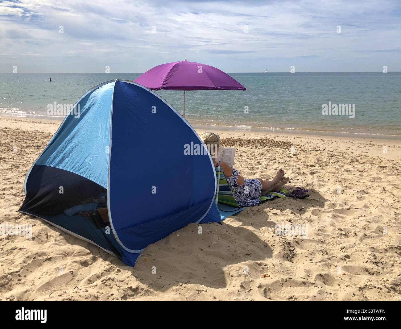 Frau, die mit einem Zelt und Sonnenschirm am Strand sitzt und ein Buch mit Blick auf das Meer liest. - Smartphone-aufgenommenes Stockfoto