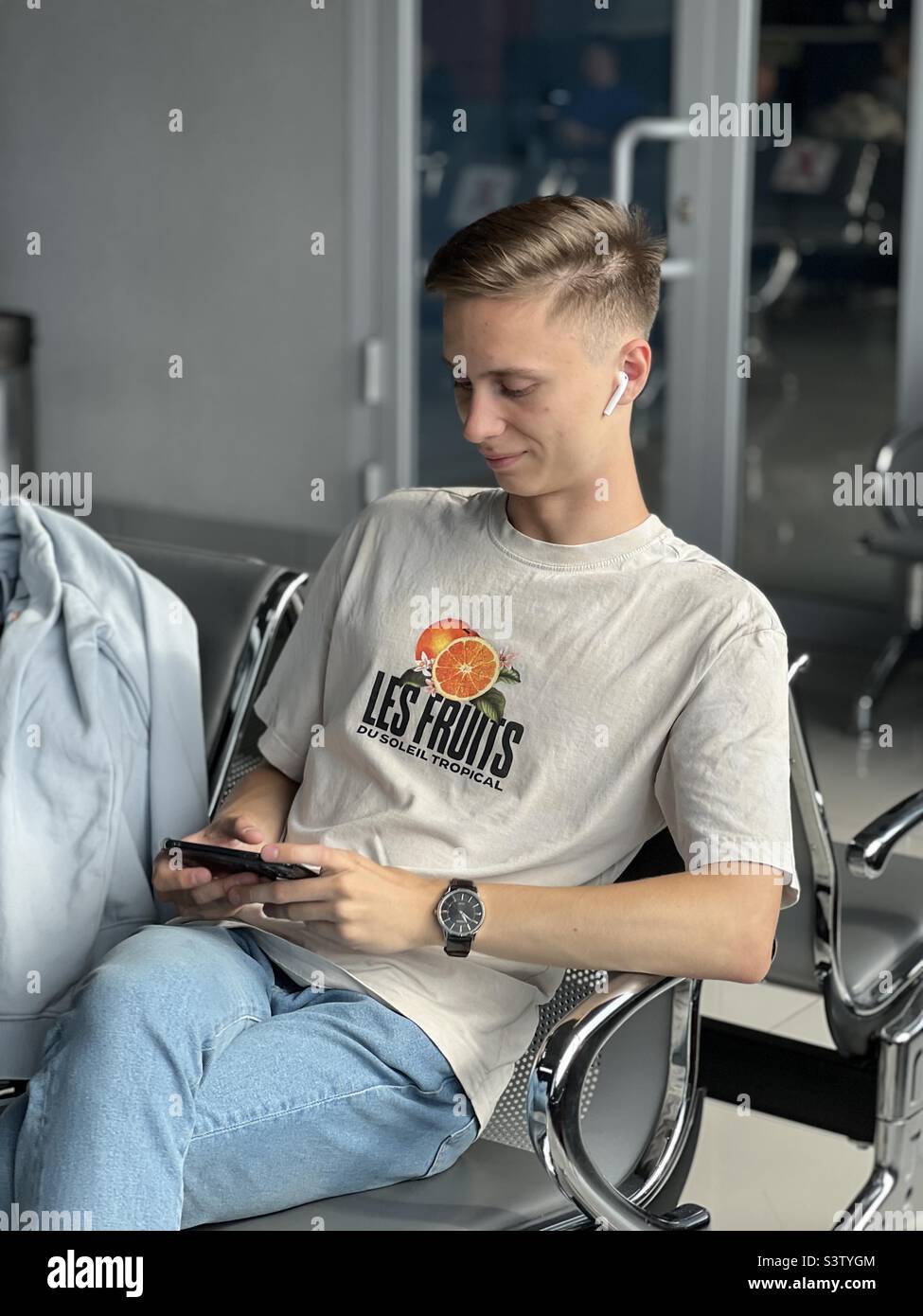 Ein junger, hübscher Kaukasier wartet auf einen Flug. Ein Mann sitzt mit einem Handy am Flughafen und schaut sich begeistert die Serie am Telefon an. Reisekonzept Stockfoto