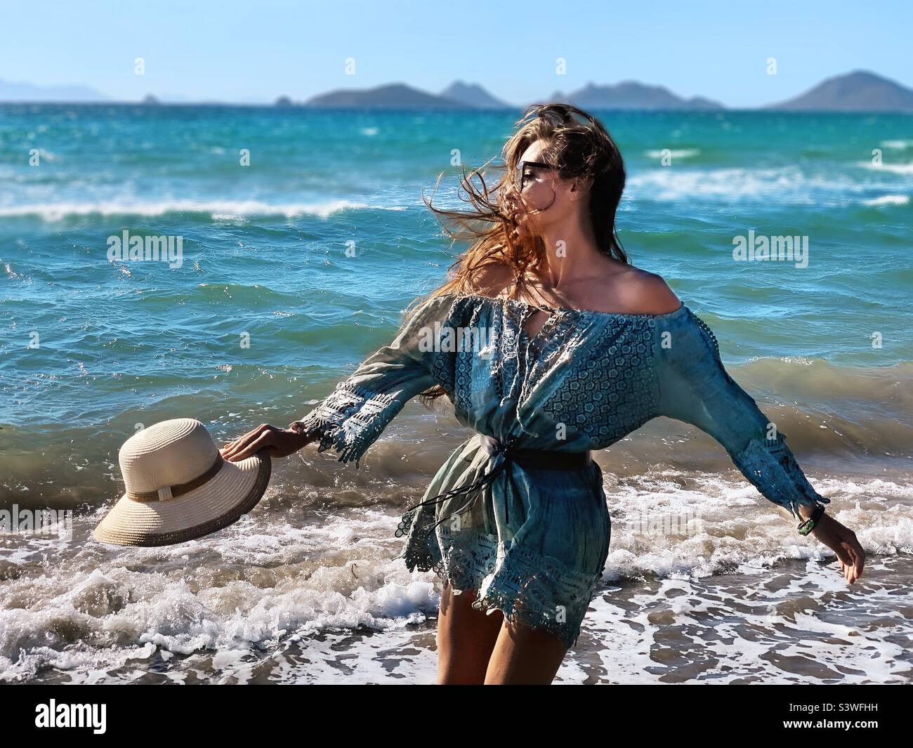 Junge Frau mit langen Haaren, die in Sommerkleidung mit Hut vor dem Meer steht Stockfoto