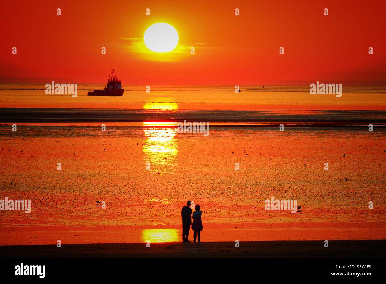 Ein wunderschöner Sonnenuntergang am Strand. Ein Boot kann direkt unter der fallenden roten Sonne gesehen werden, ein Paar blickt auf den Sonnenuntergang. Dieses Foto wurde während des außergewöhnlich heißen Wetters und einer Hitzewelle aufgenommen. Stockfoto