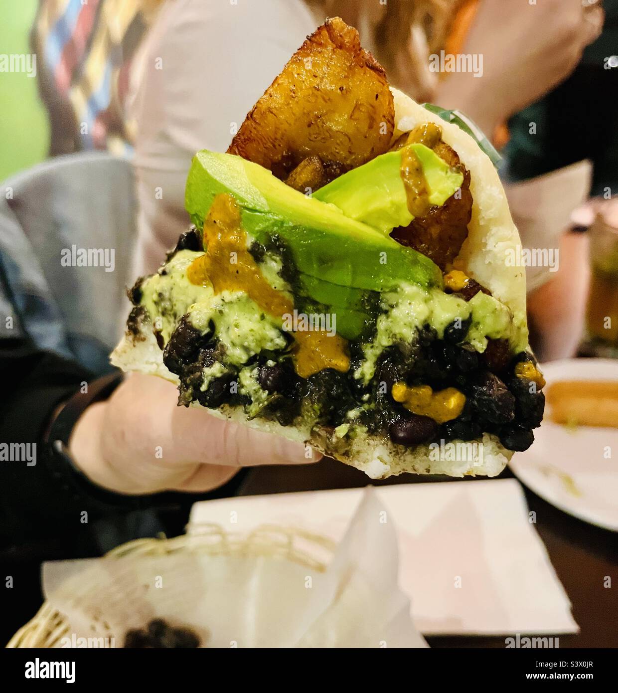 Venezolanischer Arepa gefüllt mit schwarzen Bohnen, Avocado und gebratenem Kochbananen. Stockfoto