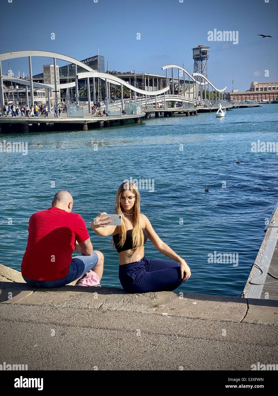 Eine Frau macht ein Selfie an der Hafenmauer am Wasser mit Blick auf die beliebte Touristenattraktion Port Vell in Barcelona Stockfoto