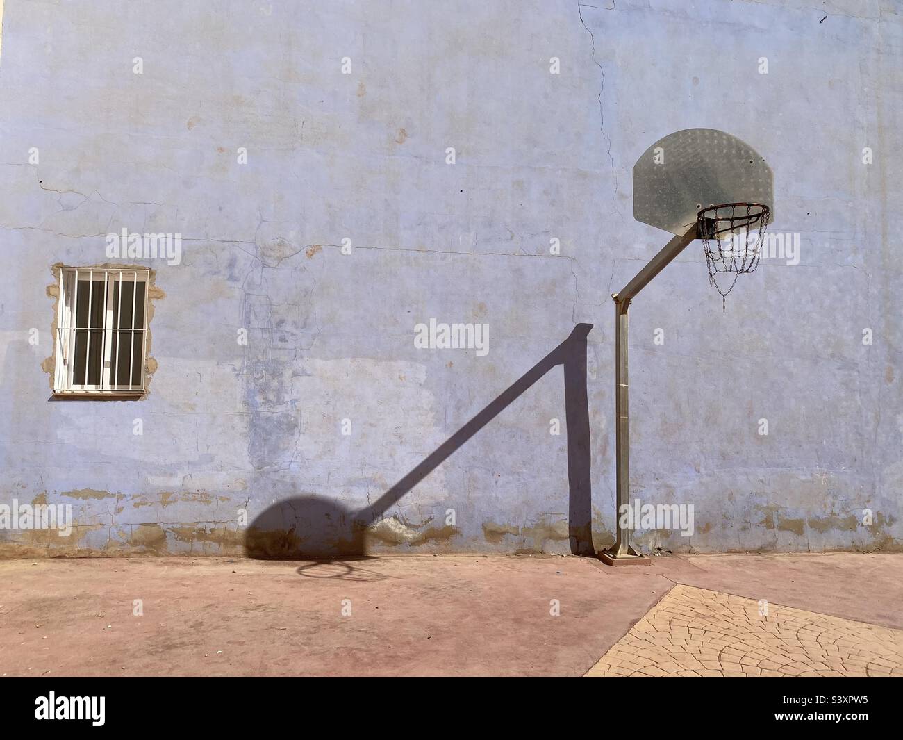 Der Schatten eines Basketballnetzes und eines Postes wird in einem scheinbar scharfen Winkel auf eine hellblaue Wand projiziert. Links befindet sich ein Fenster, das durch einen Grill geschützt ist. Stockfoto