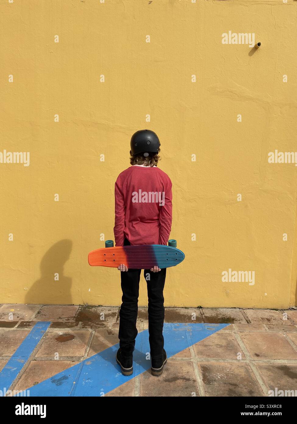 Skateboarding hält Skateboard hinter dem Rücken und blickt auf eine orangefarbene Wand Stockfoto