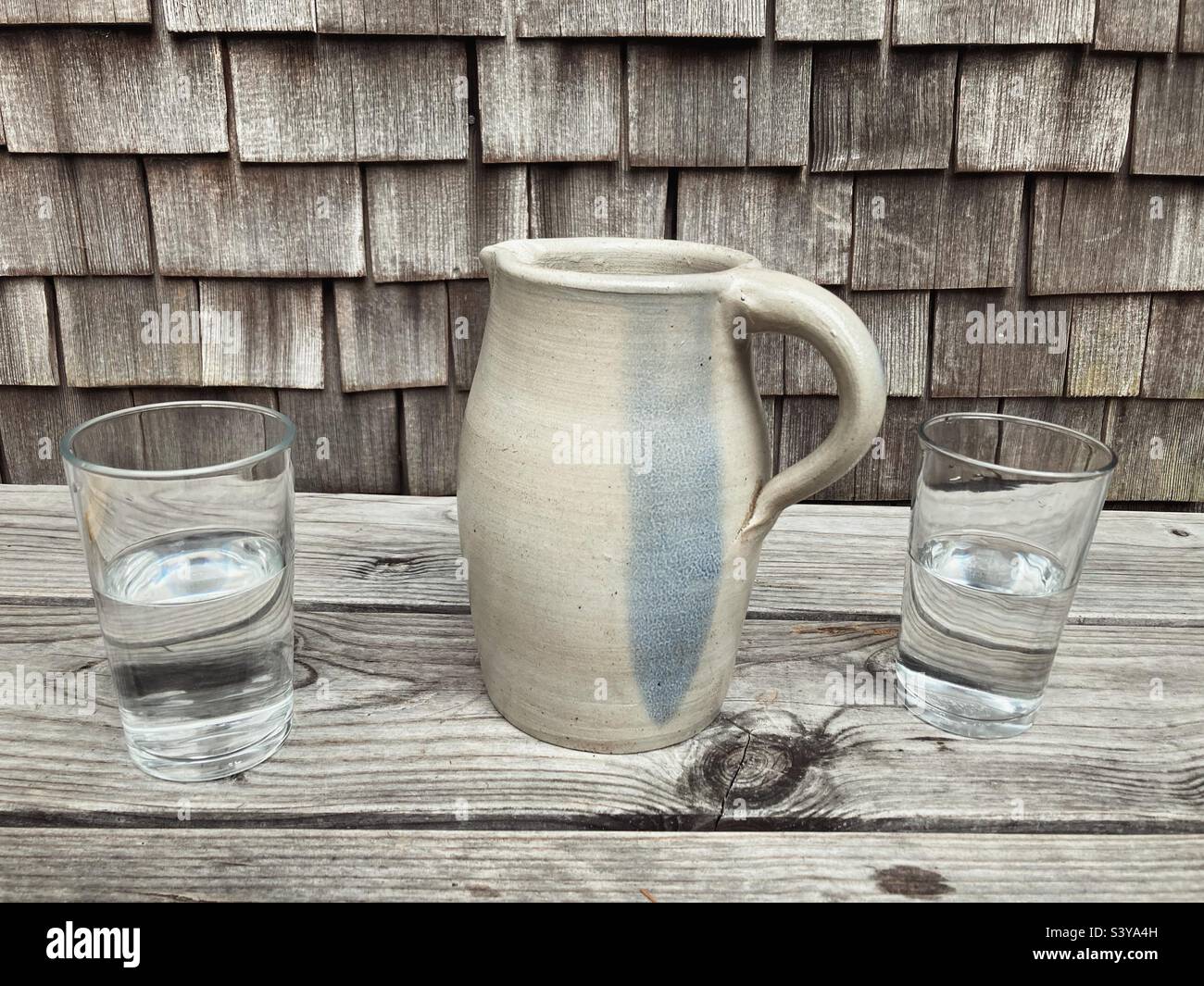 Zwei Gläser Wasser und eine Töpferwasserkanne auf einer Holzoberfläche vor einer Wand aus traditionellen Schindeln - Smartphone-aufgenommenes Stockfoto