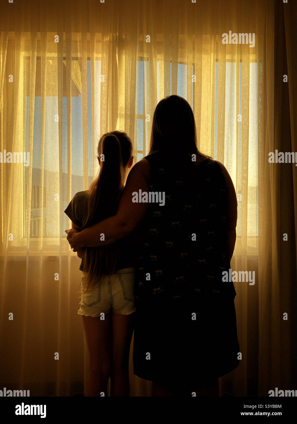 Eine Mutter umarmt ihre Tochter, während sie beide nach draußen auf den Sonnenuntergang schauen. Ein warmes Bild mit Gefühlen und eine Illustration für viele Konzepte? Foto ©️ COLIN HOSKINS. Stockfoto