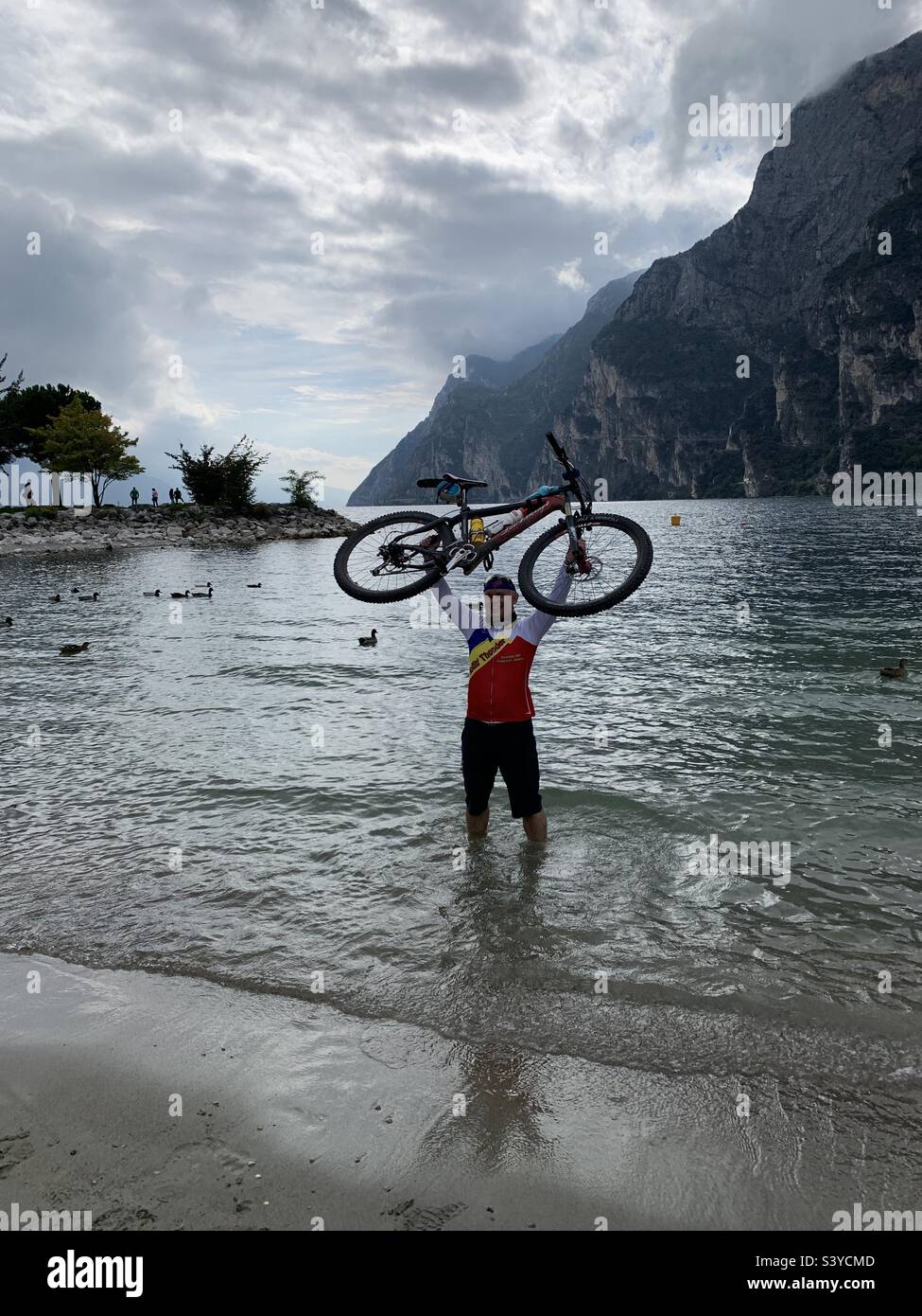 Mann, der am Gardasee Fahrrad in der Luft hält - Smartphone-aufgenommenes Stockfoto