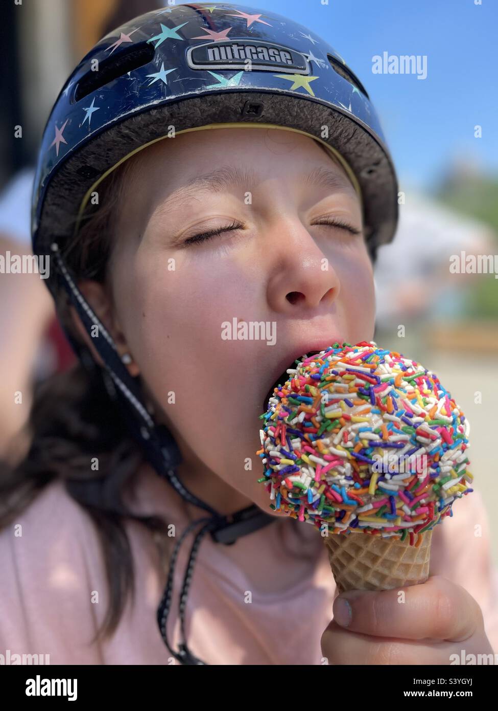 Das kleine Mädchen trägt einen Fahrradhelm und genießt einen Eiskegel, der mit bunten Prise bedeckt ist. Stockfoto