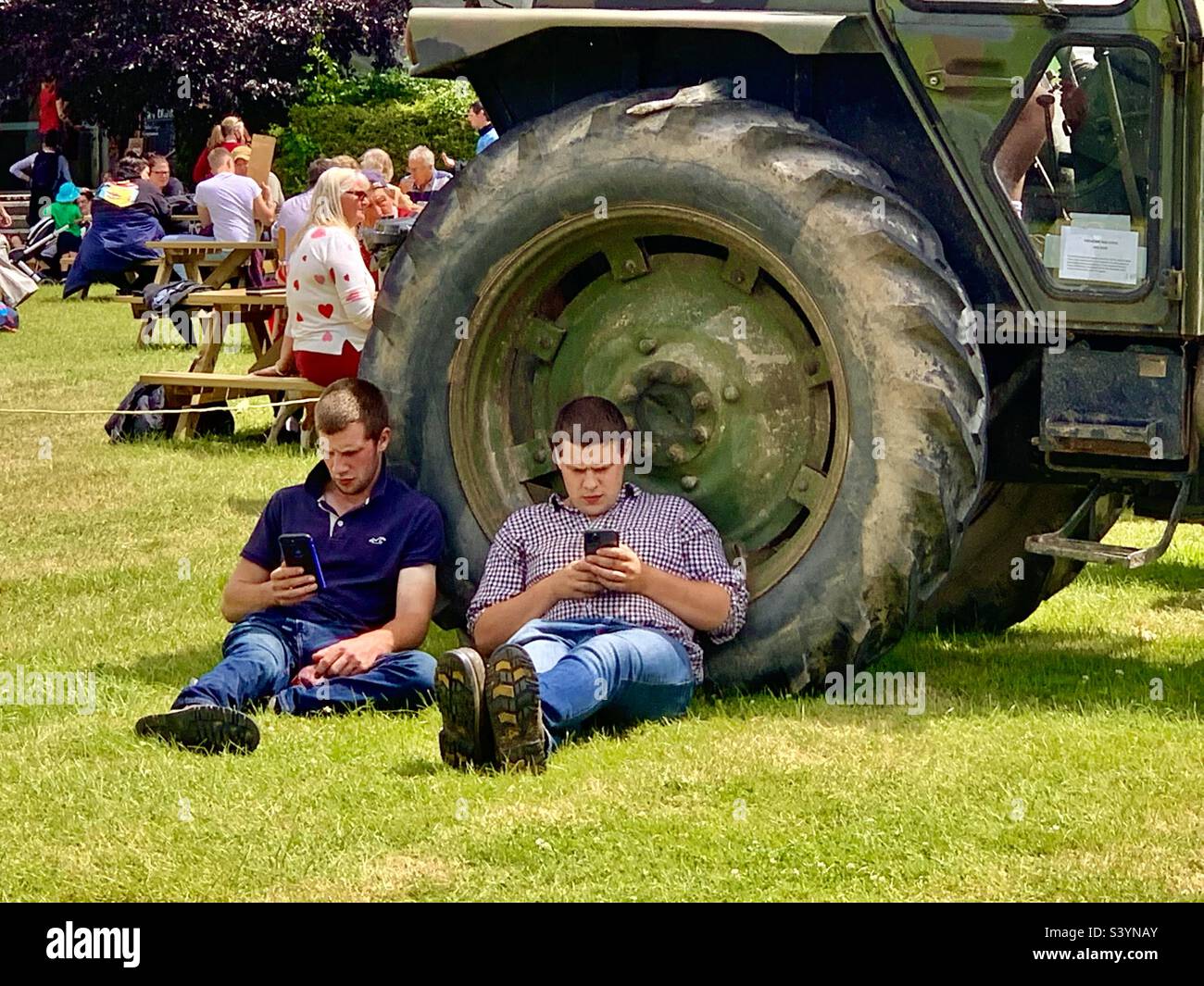 Zwei junge Bauern sitzen und entspannen sich im Schatten eines riesigen Traktorrads und nutzen soziale Medien, zwei junge Männer schreiben während der Royal Bath and West Show im Sommer in England SMS Stockfoto