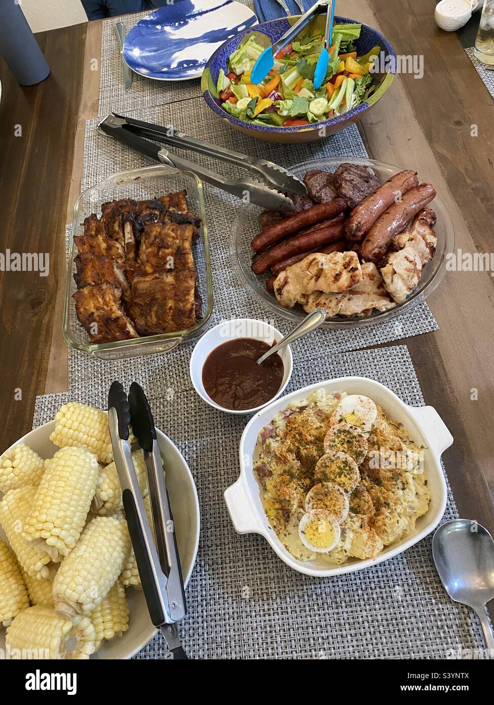 Auf dem Tisch werden verschiedene amerikanische Grillgerichte gezeigt: Hühnchen, Hotdogs, Hamburger, Steak, Maiskolben, Bohnen, Rippchen, Ei- und Kartoffelsalat und Salat. - Smartphone-aufgenommenes Stockfoto