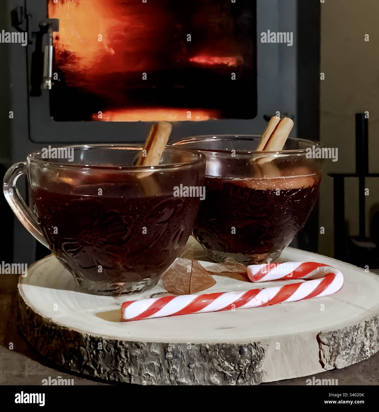 2 Punchgläser voller Glühwein auf einer Holzplatte mit Zuckerrohr vor einem brüllenden Holzbrenner. Stockfoto