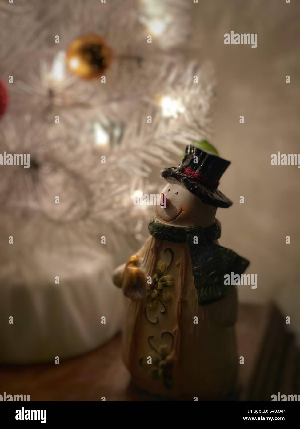 Glückliche Schneemannfigur aus Keramik unter einem weißen Weihnachtsbaum, beleuchtet mit weißen Lichtern - Smartphone-aufgenommenes Stockfoto