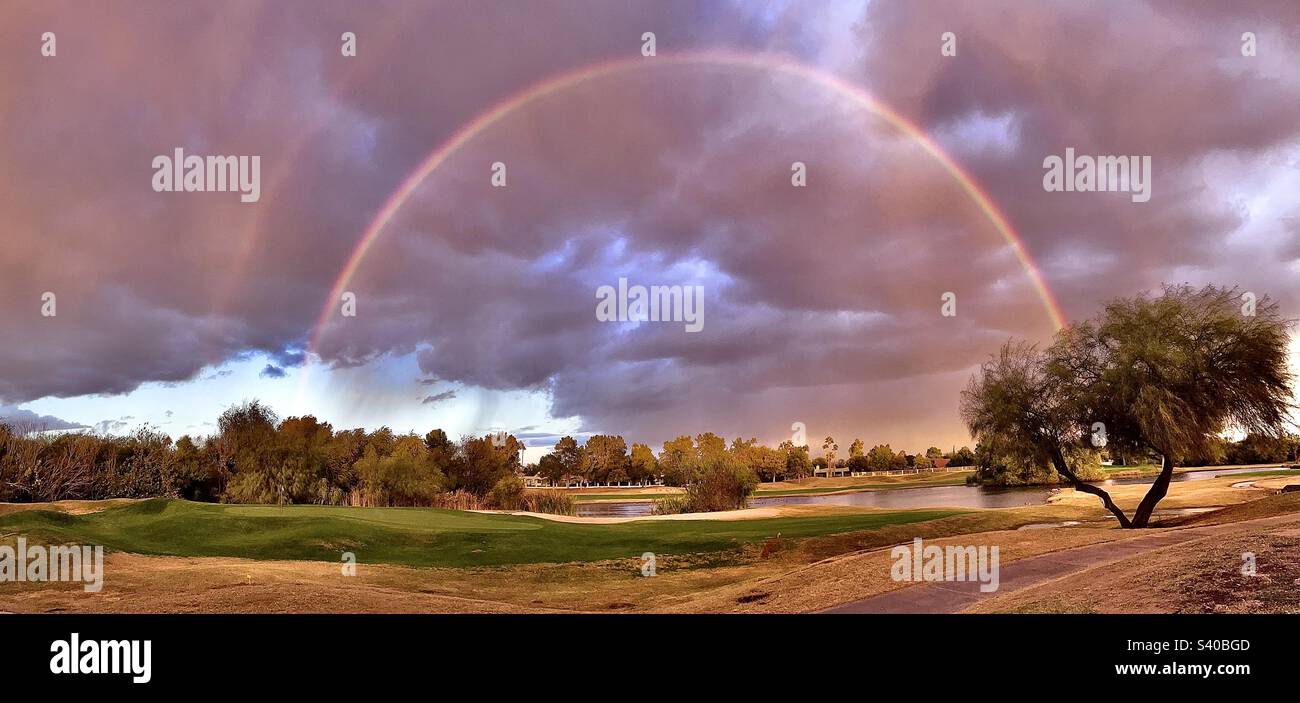 Ein Regenbogen über einem Teich mit Golfwagen und einem Baum mit verzweigten Stämmen im Vordergrund - ein Hauch eines doppelten Regenbogens gegen stürmischen Himmel, der von der untergehenden Sonne bemalt wird Stockfoto