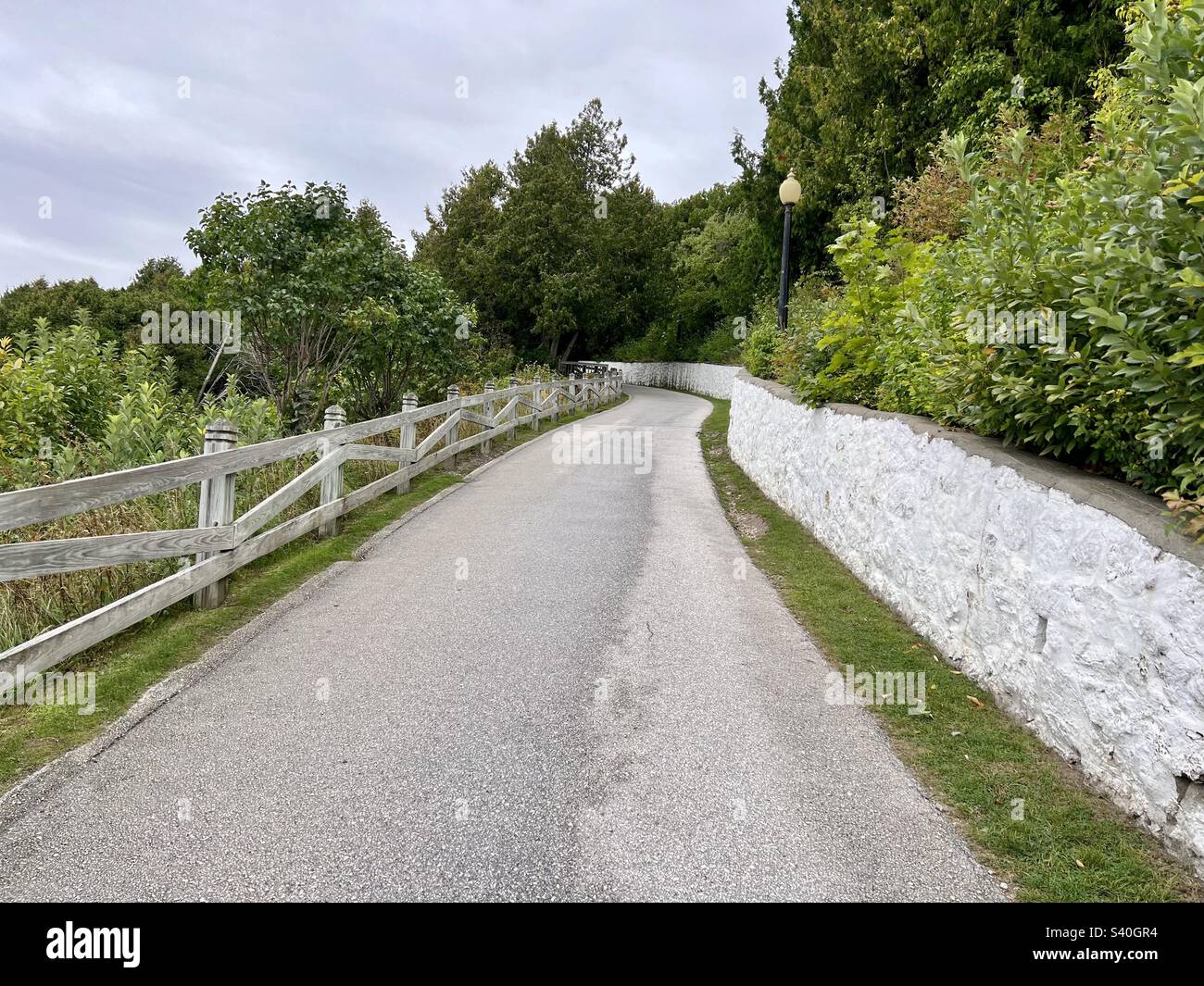 Gepflasterte Straße, die einen Hügel hinaufführt, mit einem weißen Zaun auf der einen Seite und einem Steinzaun auf der anderen. Wunderschöne grüne Bäume und Sträucher säumen die Straße. Foto aufgenommen auf Mackinac Island Michigan Stockfoto