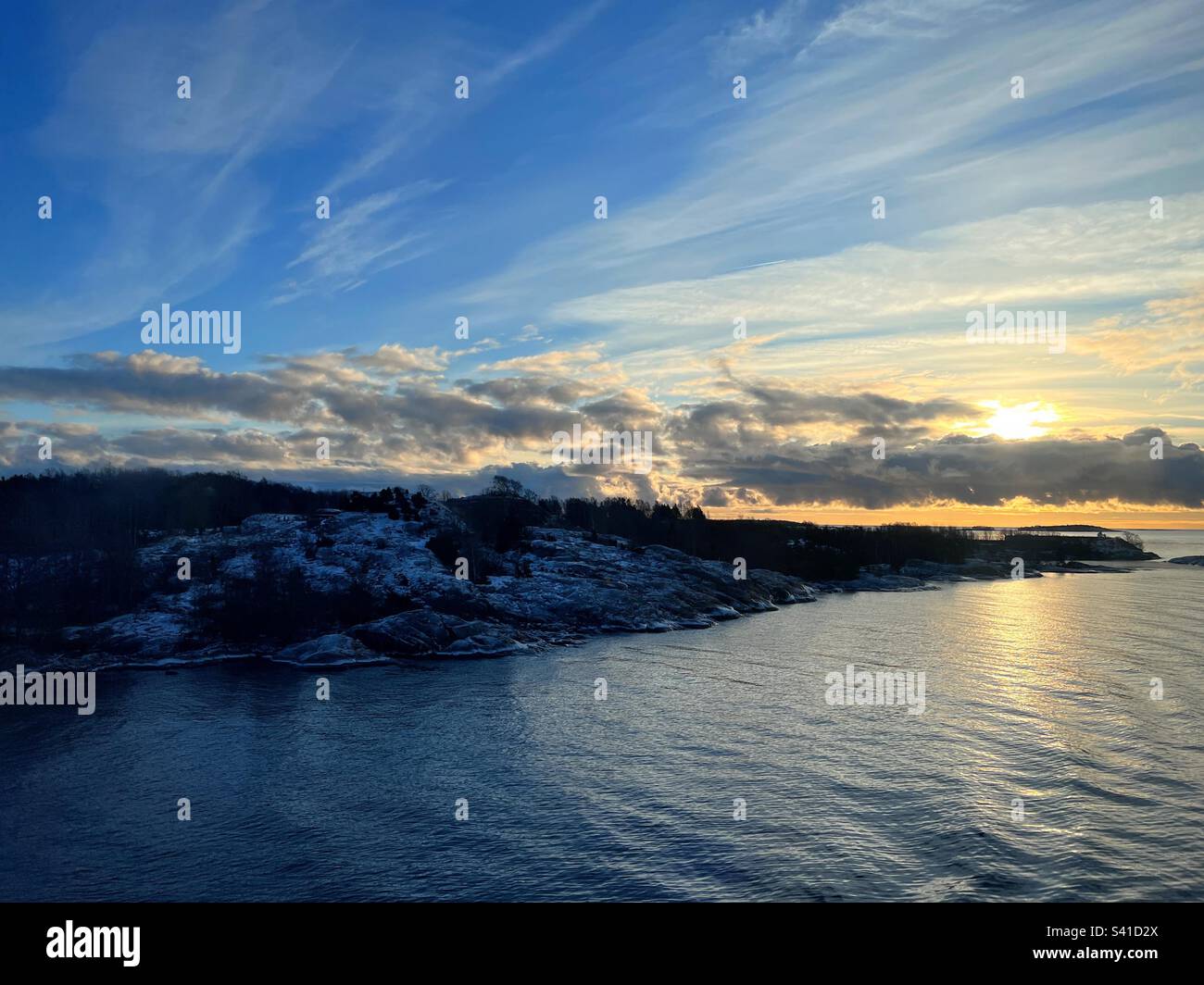 Ruhige Morgenlandschaft mit dunklem Landpeninsula und Sonnenaufgang hinter niedrigen Wolken mit Reflexion im Meerwasser Stockfoto