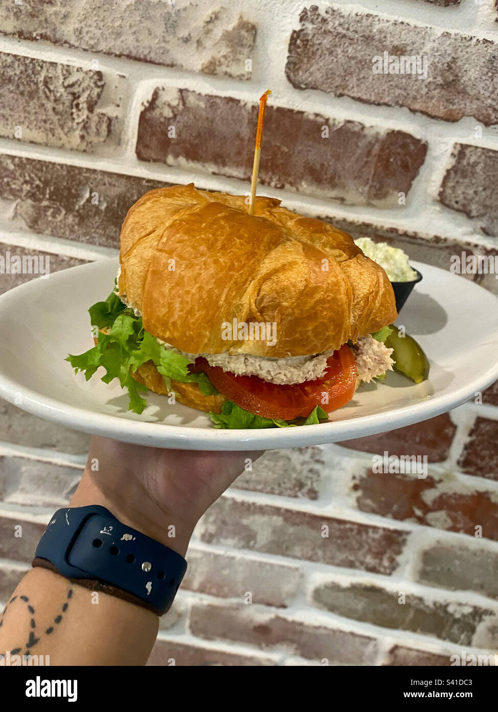 Thunfischsalat-Sandwich auf einem Croissant mit Salat und Tomaten. Serviert auf einem weißen Teller. Backsteinwand im Hintergrund. Angewinkeltes Foto. Stockfoto