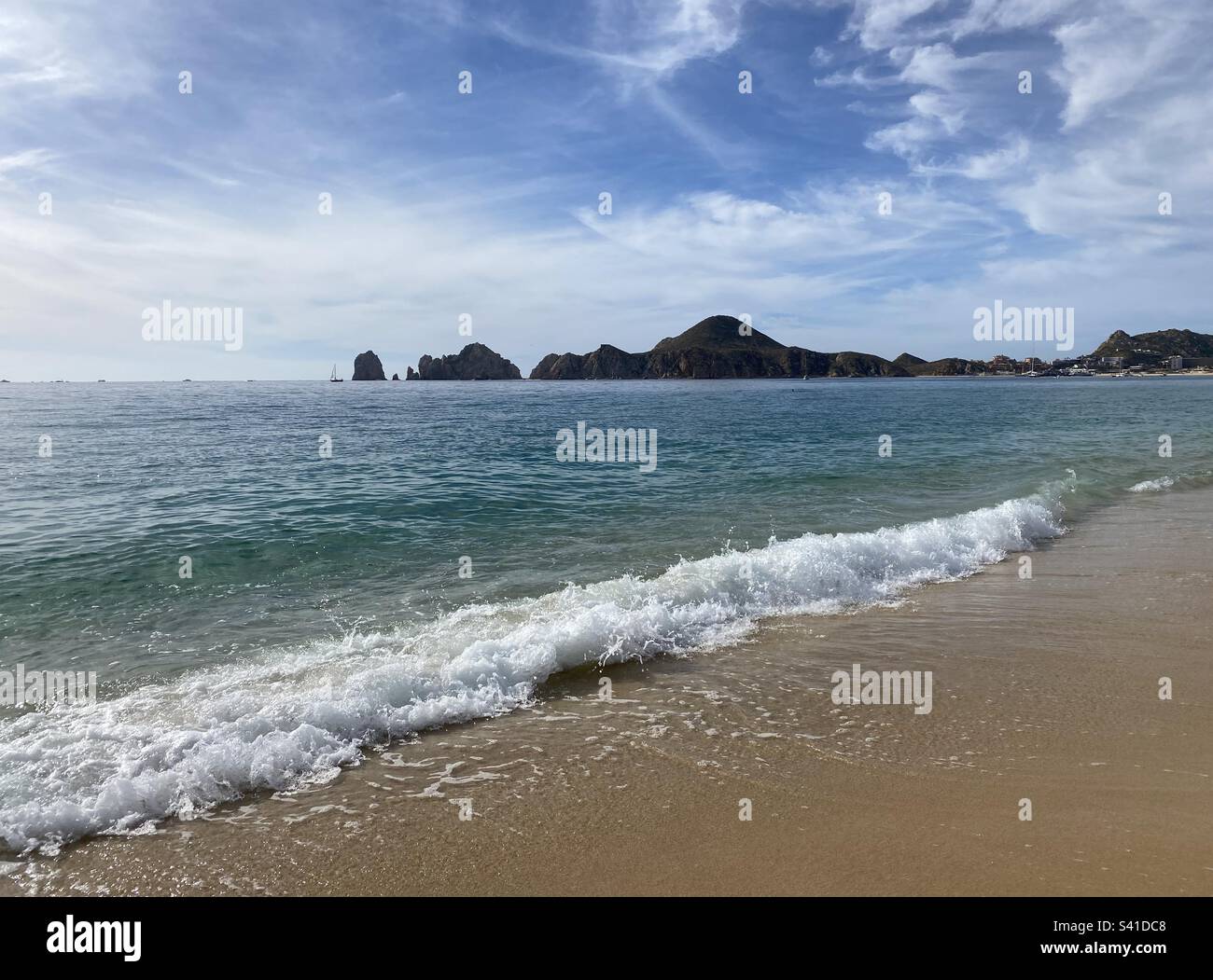 Die Welle stürzt auf einen Strand mit Felsformationen im Hintergrund. Blauer Himmel mit ein paar Wolken. Stockfoto