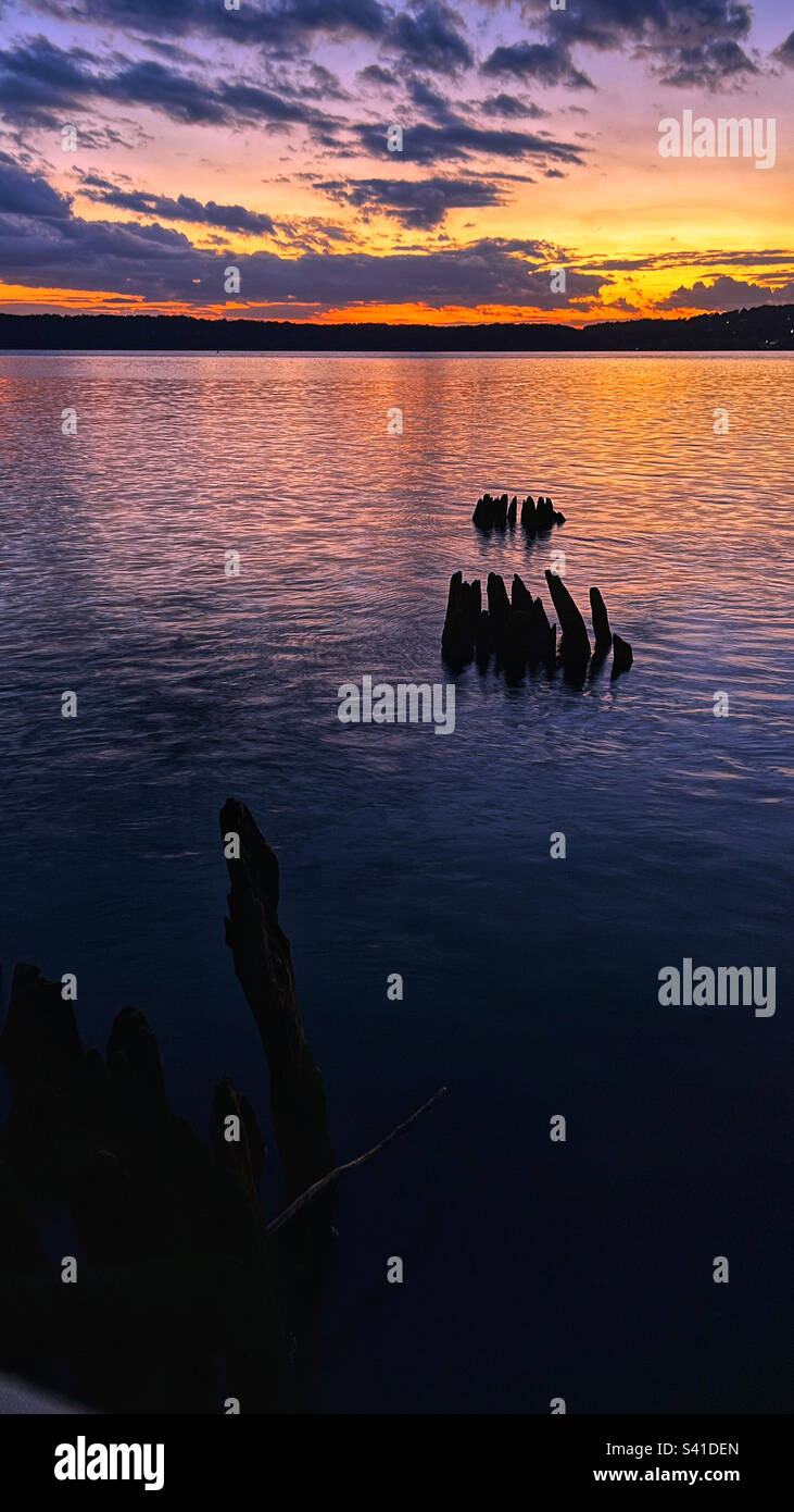 Vertikales Bild mit Blick über den Hudson River in Richtung spektakulärer violetter, orangefarbener und magentafarbener Sonnenuntergänge mit Silhouette vom zerstörten alten Dock. - Smartphone-aufgenommenes Stockfoto