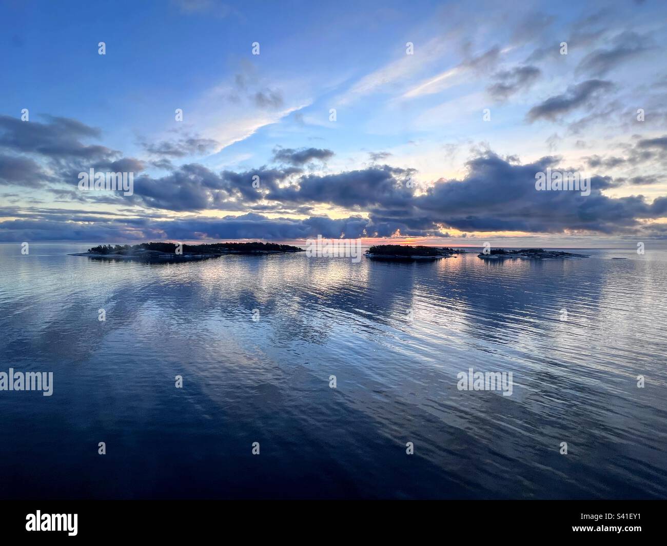 Ruhige Szenenperspektive über dem Sonnenaufgang im Meer hinter dunklen Wolken mit kleiner Insel und Reflexion im Wasser Stockfoto