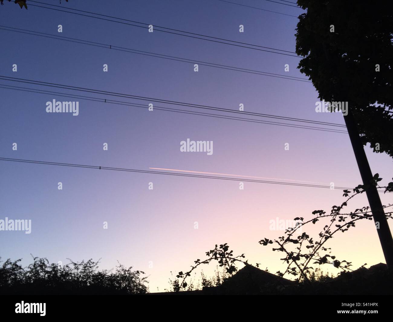Abendhimmel mit Silhouetten, Dächern und Stromleitungen. Die weiße Spur eines Düsenflugzeugs scheint parallel zu einem der elektrischen Kabel zu fliegen. - Smartphone-aufgenommenes Stockfoto