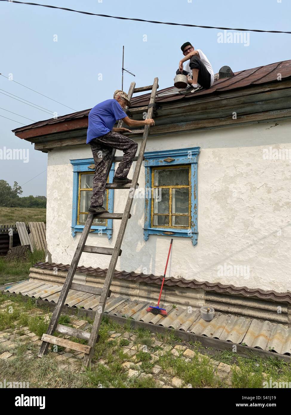 Ein älterer Mann, der sich gebeugt hat, klettert die Treppe zum Dach eines Dorfhauses, um seinem kleinen Sohn mit einer Dose Farbe zu helfen. - Smartphone-aufgenommenes Stockfoto