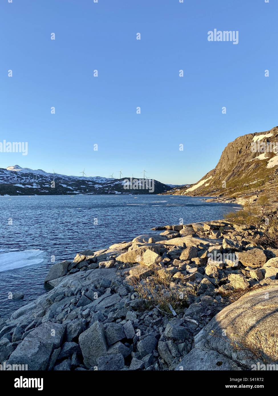 Nedre Jernvatnet bei Narvik, Norwegen, an einem sonnigen Sommerabend. Es schwimmt immer noch etwas Eis im Wasser, aber die Sonne scheint und das Wetter ist gut. Sie können Windgeneratoren in der Ferne sehen. - Smartphone-aufgenommenes Stockfoto