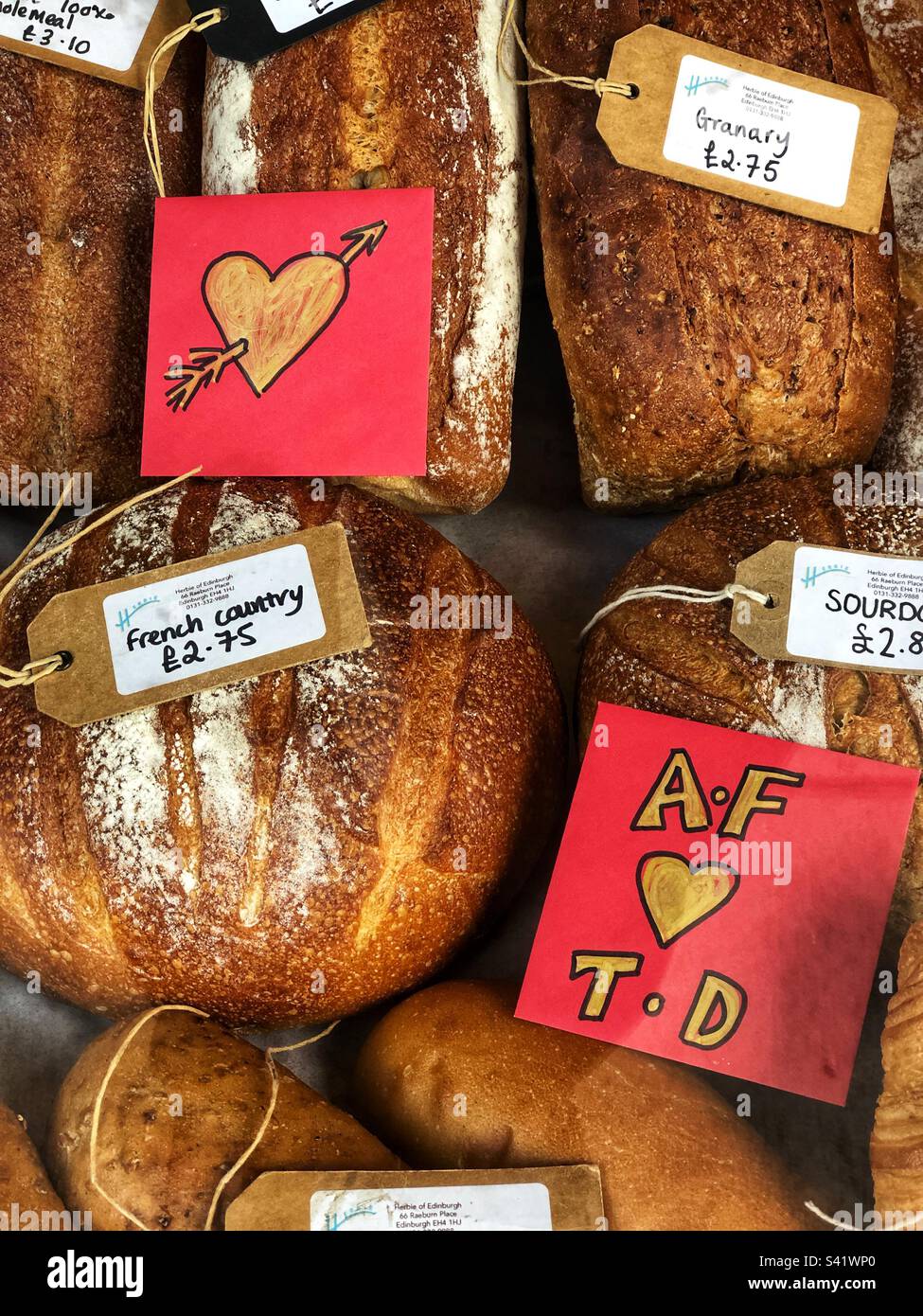 Frische, handwerklich gekrustete Brotlaibe mit Preisschildern in einem Delikatessengeschäft und einer Schaufensterausstellung für Wurstwaren kurz vor dem Valentinstag - Smartphone-aufgenommenes Stockfoto